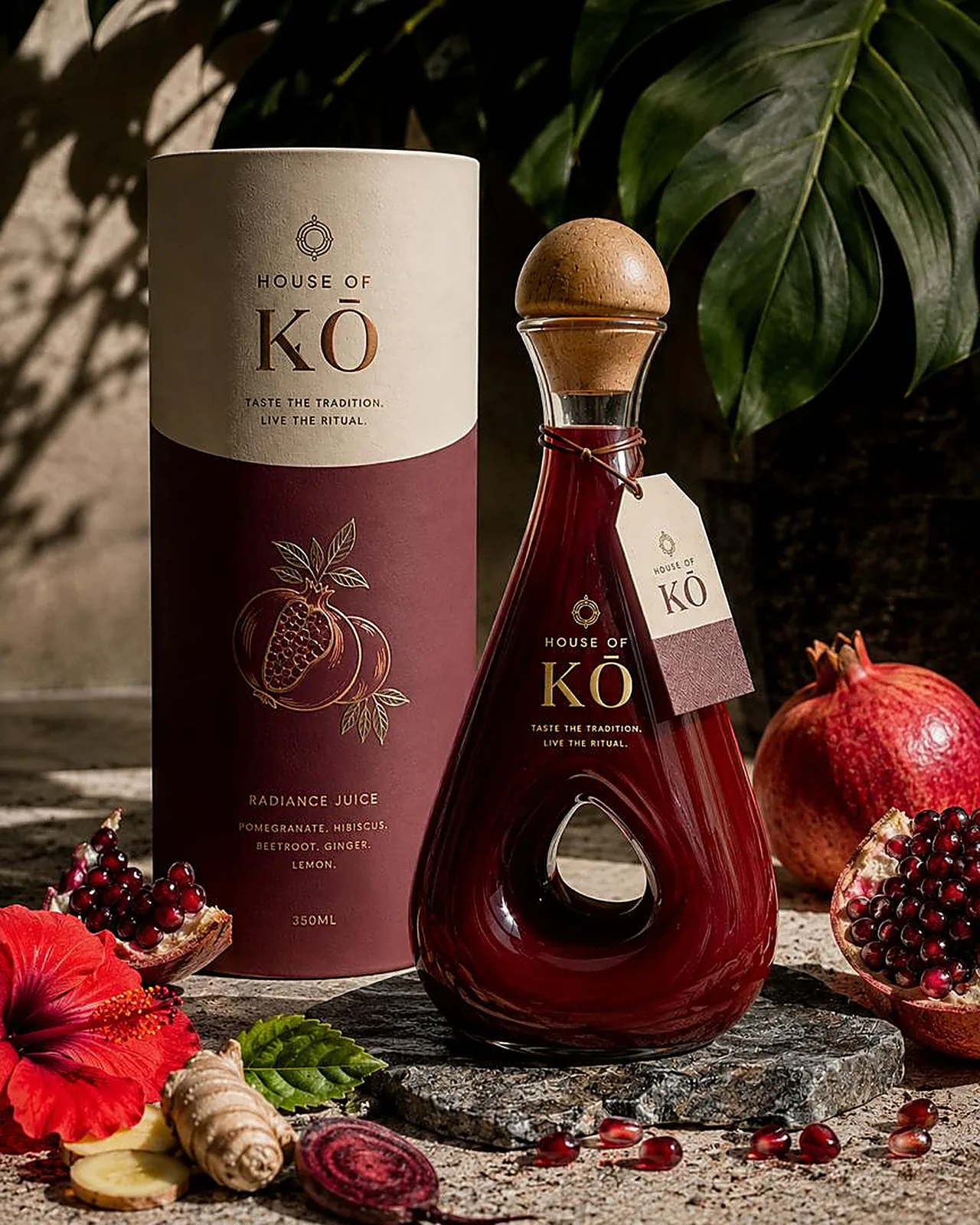 House of KŌ Radiance Juice in the signature teardrop glass bottle with wooden stopper and neck tag, beside its burgundy gift tube with fresh pomegranate, hibiscus and beetroot
